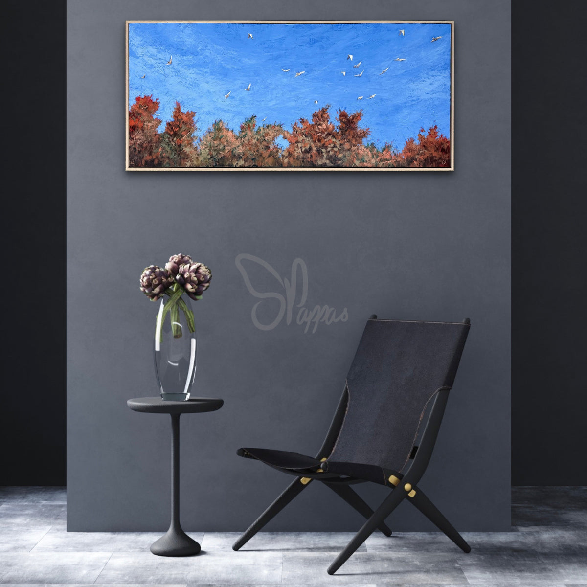 A framed horizontal canvas print of a blue sky with flying birds and a line of red and orange autumn trees, displayed on a dark gray wall above a black lounge chair and a small side table with flowers.